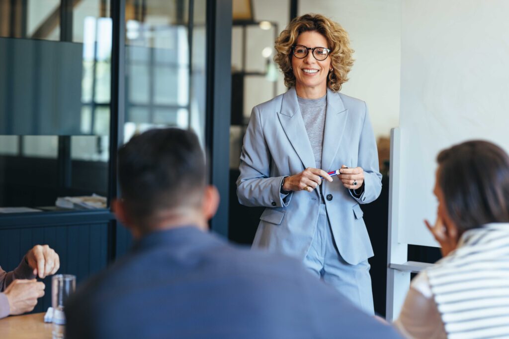 A lady in a suit stands whilst in front of a couple of work colleagues in a work meeting