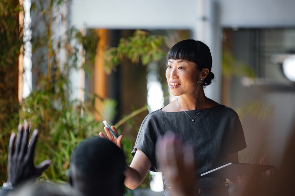 A confident presenter addresses an audience during a business seminar, highlighting her engaging speech. The enthusiastic participants raise their hands for questions.
