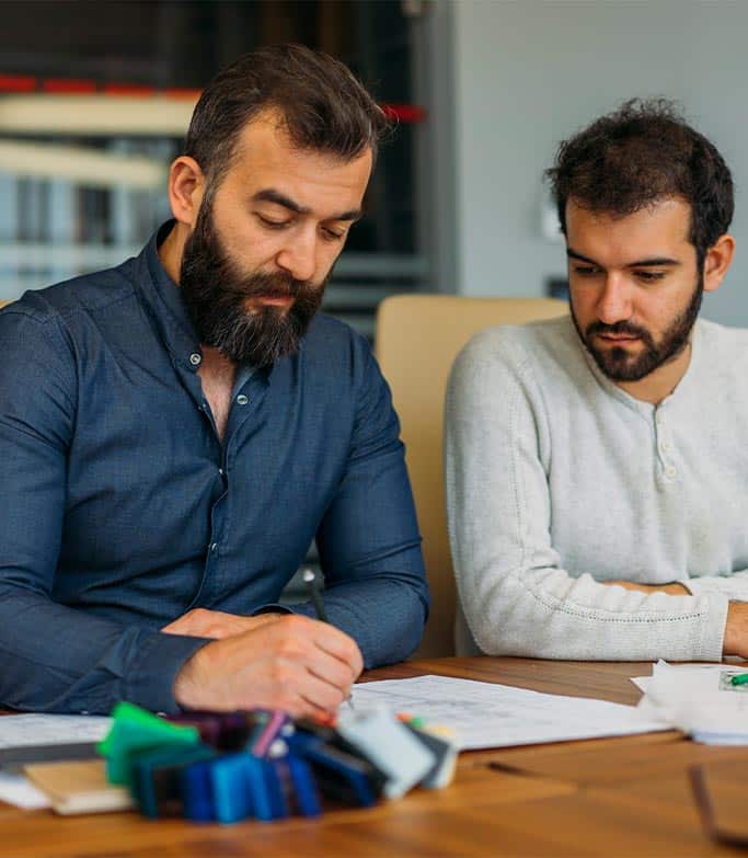 Two men sit together at a desk in front of a shortlist / paperwork
