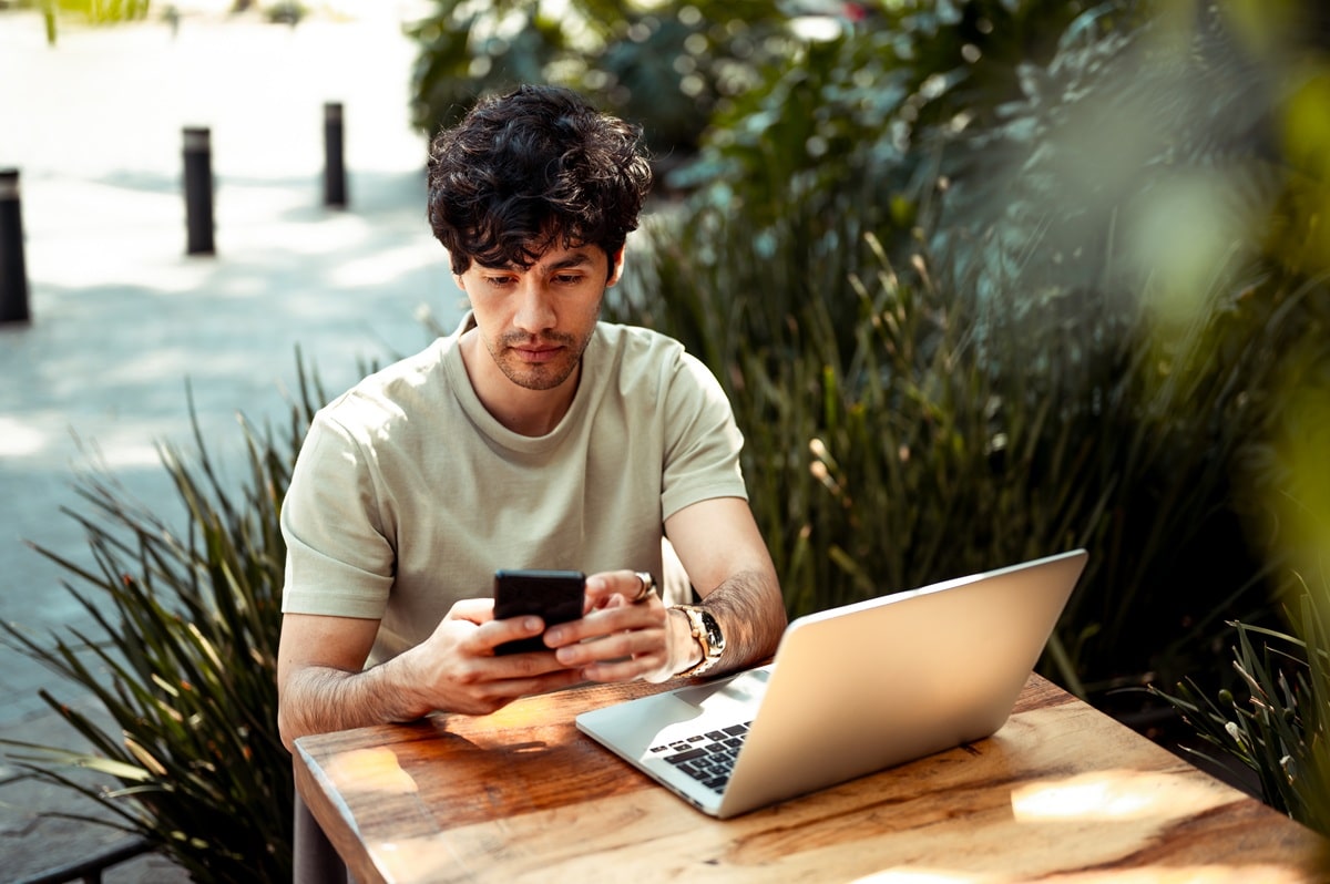 Man working from coffee shop. He is using mobile phone and laptop